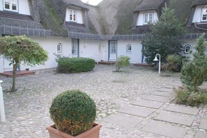 A courtyard with stone paving, a round potted plant, and a building with a thatched roof. - "Syltperle" in the Sölring-Hüs (Sylt\/Rantum)