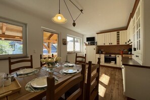 Un coin repas avec une table en bois dressée pour quatre personnes, un luminaire suspendu et une cuisine dotée de meubles blancs et d’un four à micro-ondes.
