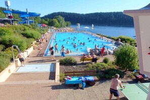 A swimming pool area with people swimming and sunbathing, surrounded by greenery and overlooking a body of water. - Holiday home in houses with swimming pool (Häusern)