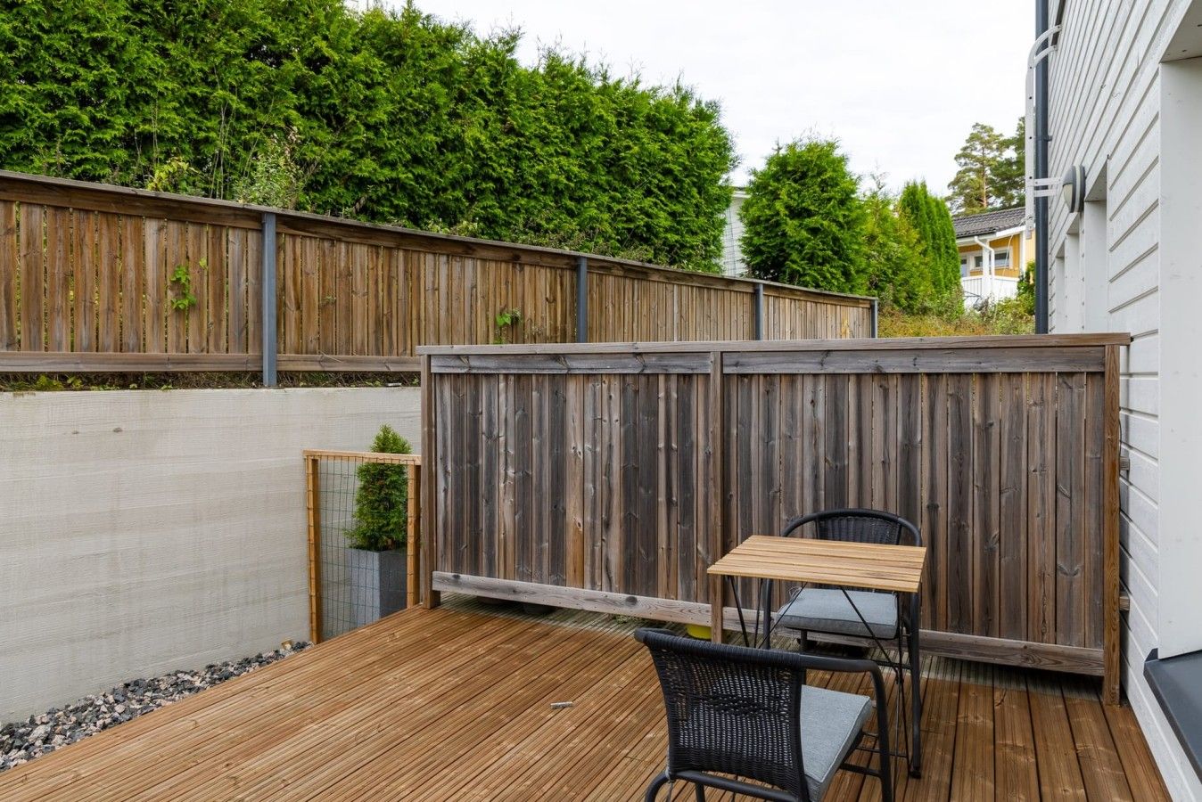 Une terrasse en bois avec une table et une chaise, entourée d’une clôture en bois et d’une haie.