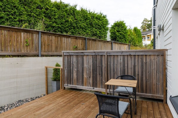 A wooden deck with a table and chair, surrounded by a wooden fence and a hedge.