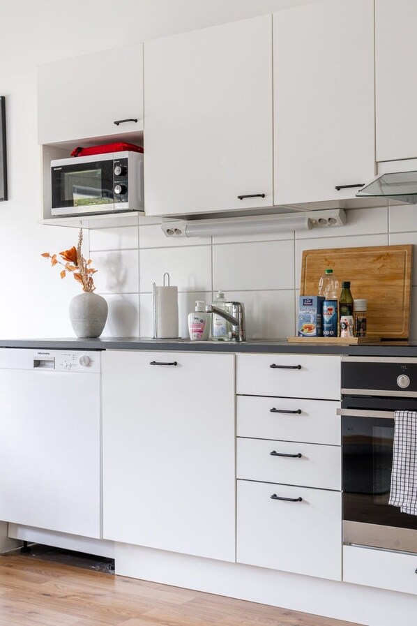 A modern kitchen with white cabinetry, a microwave, a dishwasher, and a wooden cutting board with bottles.