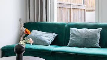 Un canapé vert agrémenté de deux coussins, une petite table d’appoint ronde en bois sur laquelle reposent un vase et une télécommande, et une vue sur une terrasse en bois et la verdure environnante.