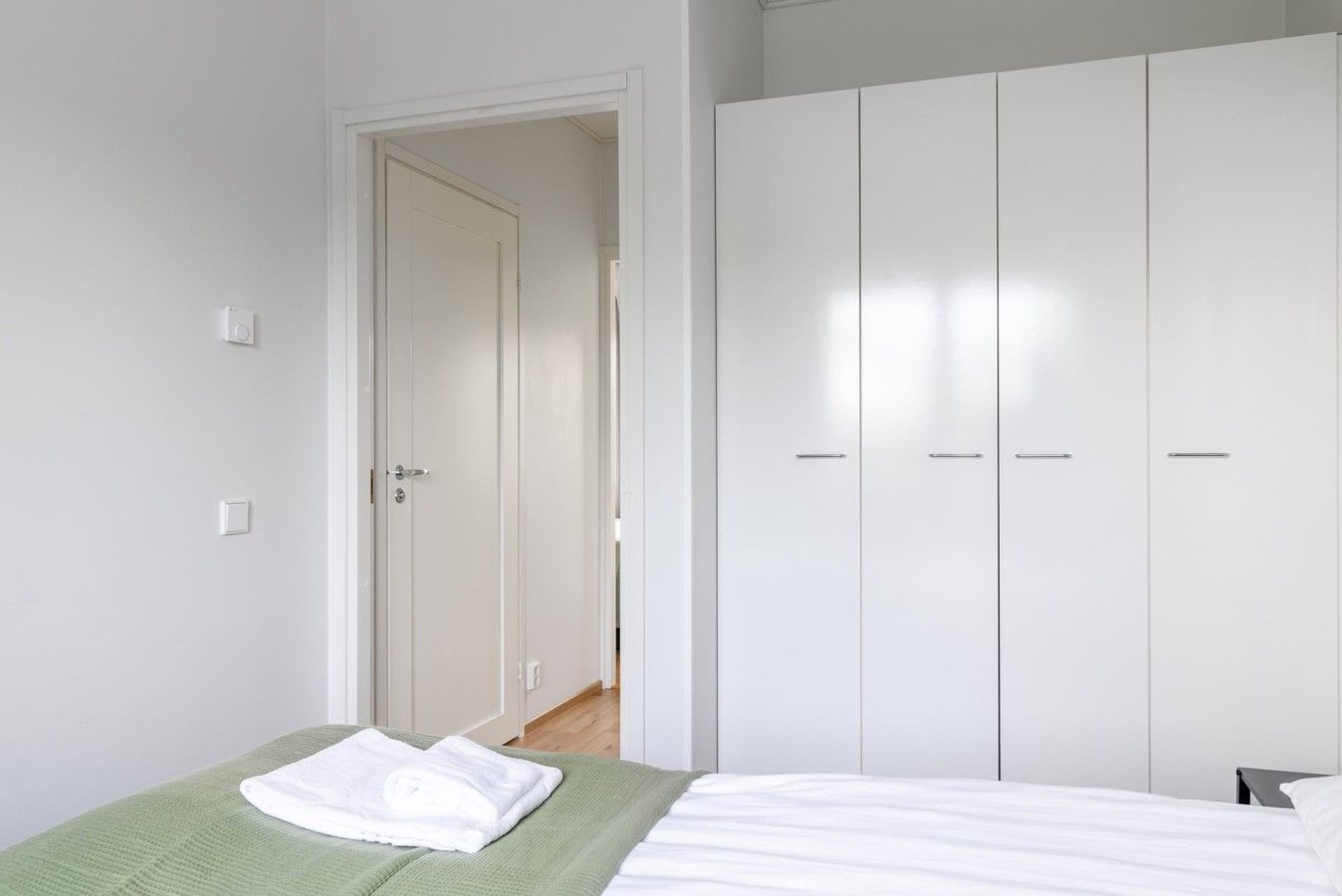 A bedroom with a bed, a green blanket, a white towel, and a white wardrobe.