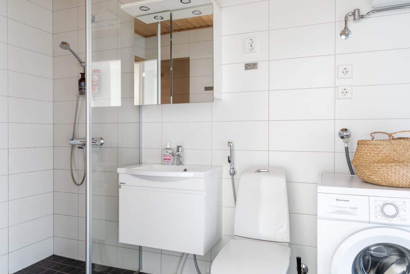 Moderni kylpyhuone, jossa on lasinen suihkuseinä, valkoinen pesuallas, wc ja pesukone.