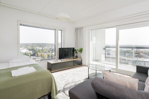 Moderni hotellihuone, jossa on sänky, sohva, televisio ja suuret ikkunat, joista avautuu näkymä kaupunkiin.