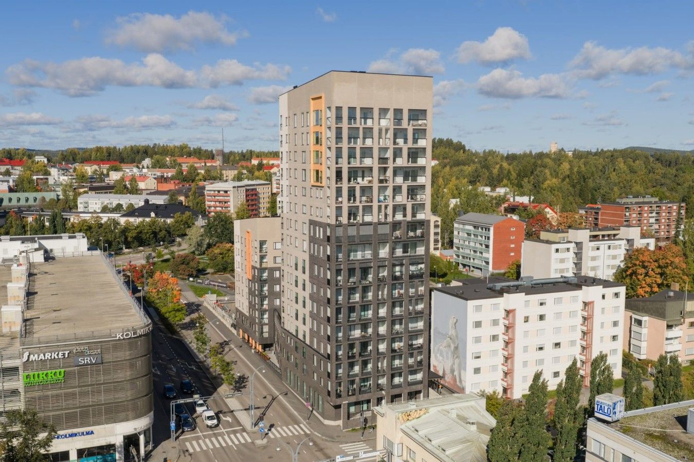 Korkea kerrostalo, jossa on useita kerroksia, ja sen ympärillä on muita rakennuksia ja puita.