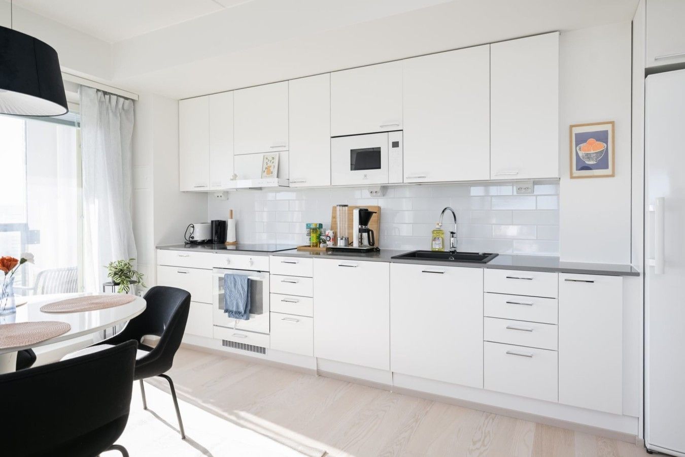 Een moderne keuken met witte kasten, een magnetron en een spoelbak. Er is een eethoek met een tafel en stoelen.