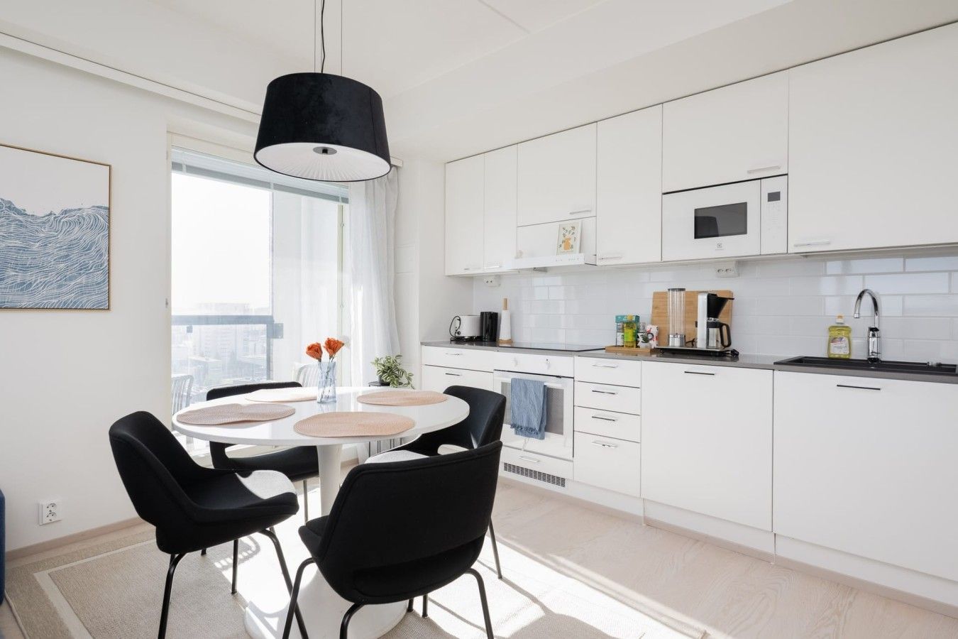 Une cuisine moderne avec des meubles blancs, un coin repas doté d’une table ronde et de chaises noires, et un luminaire suspendu.
