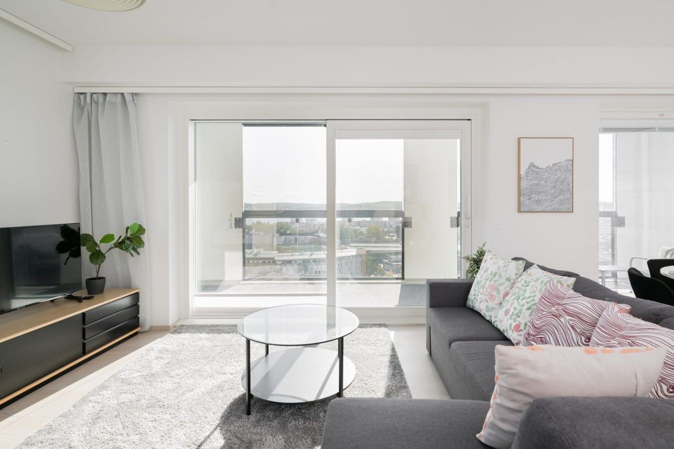 Un soggiorno moderno con una TV a schermo piatto, un tavolino in vetro, un divano grigio e un'ampia porta scorrevole in vetro che dà su un balcone con vista sulla città.