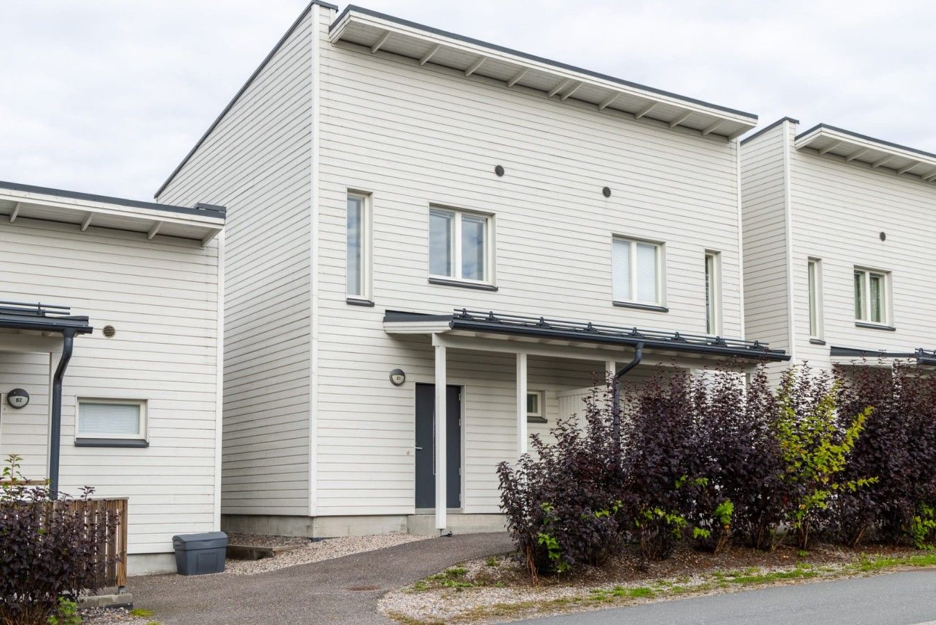 Moderni, kaksikerroksinen asuintalo, jossa on tasakatto, katettu kuisti ja pieni etupiha, jossa on pensaita.