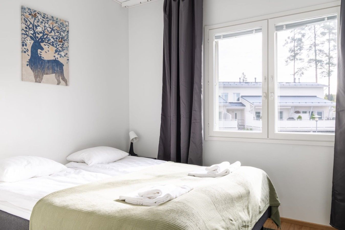 A hotel room with a bed, a window with a view of buildings, a painting on the wall, and a lamp.