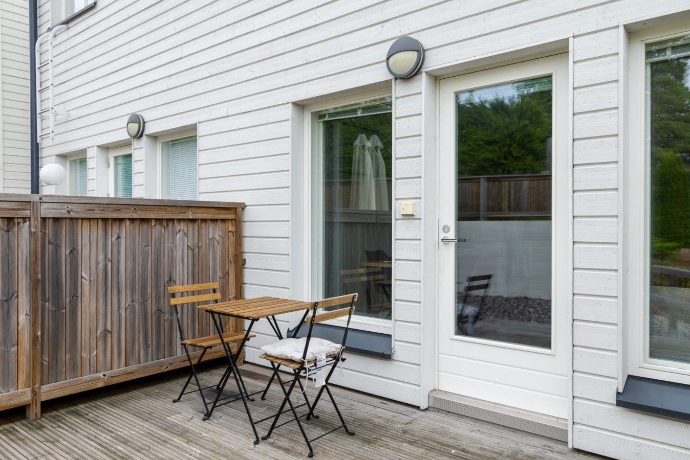 A wooden deck with a table and chairs, a wooden fence, and a house with white siding and large glass doors.