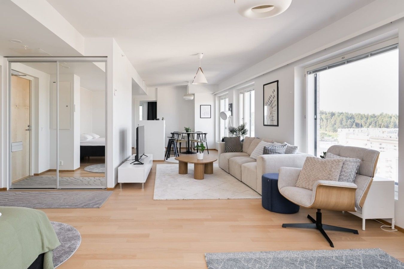 Una sala moderna con un sofá, una mesa de centro y una televisión. Hay una zona de comedor con sillas y una mesa. La habitación tiene ventanas grandes y vistas al exterior.