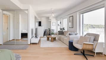 Una sala moderna con un sofá, una mesa de centro y una televisión. Hay una zona de comedor con sillas y una mesa. La habitación tiene ventanas grandes y vistas al exterior.