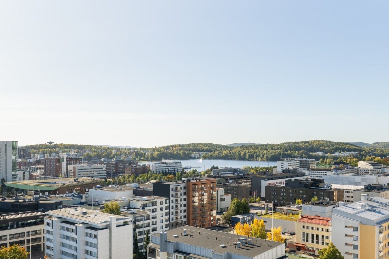 Kaupunkikuva, jossa on moderneja rakennuksia, vesialue ja taustalla metsäinen kukkula.