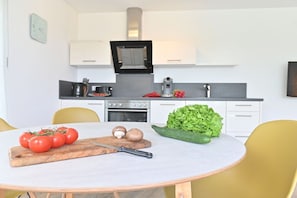Eine moderne Küche mit weißen Schränken, einem Herd und einem Spülbecken. Ein Tisch zeigt ein Schneidebrett, Tomaten und ein Messer.