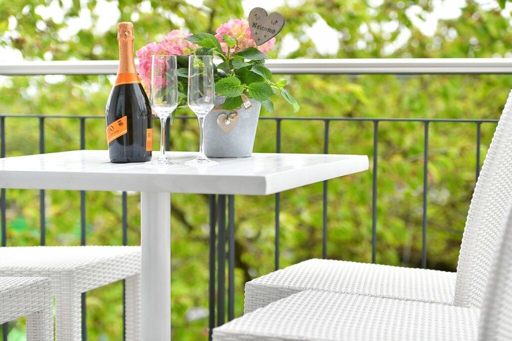 Een witte tafel met een champagnefles, twee glazen en een bloemstuk op een balkon met een metalen reling.