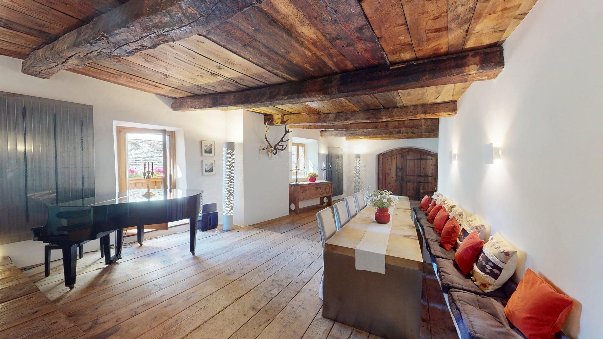 Ein geräumiger Raum mit Holzbalkendecke, einem Klavier, einem langen Esstisch mit Stühlen und einer Couch mit Kissen.