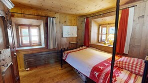 Ein Himmelsbett mit einem rot-weißen Musterüberwurf, Holzwänden und einem Fenster mit roten Vorhängen.