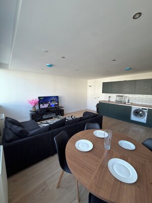 A modern living room with a black sofa, a wooden dining table with plates, and a TV displaying a show. - Modern 3 Bedroom Flat on Eltham High Street| 7 mins from the Station! (London)