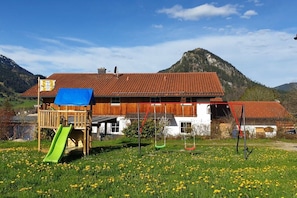 Un bâtiment en bois surmonté d’un toit de tuiles rouges, une terrasse en bois avec un toboggan vert, et une chaîne de montagnes en arrière-plan.