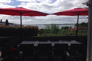 An outdoor patio with a table and chairs, red umbrellas, and a view of a body of water. - Holiday home with 4 bedrooms (Egernsund)