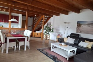 A spacious living area with a wooden ceiling, a dining table with chairs, a black leather sofa, and a staircase leading to an upper level. - Holiday home in Hooksiel with swimming pool (Hooksiel)