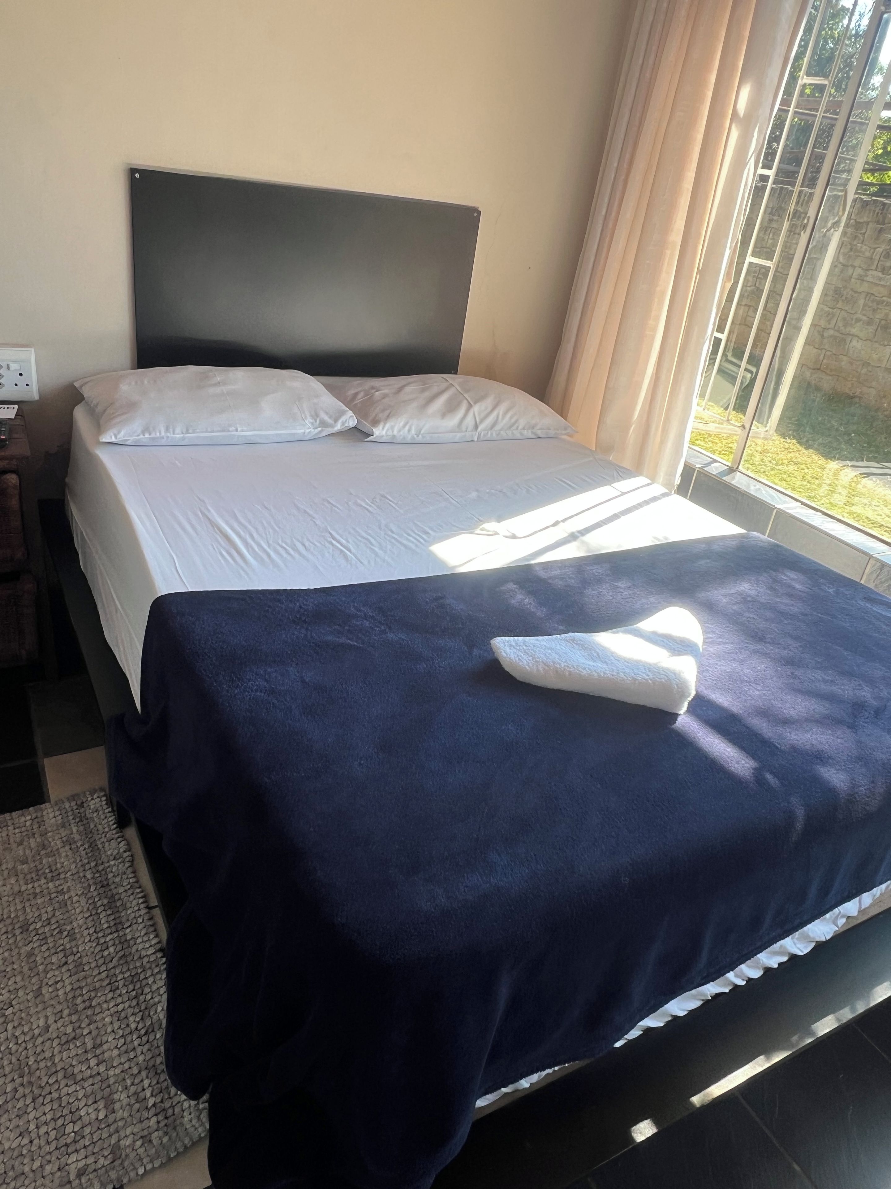 Un lit recouvert d’une couvre-lit bleu foncé et orné d’oreillers blancs, une table de chevet avec une lampe et une fenêtre avec des rideaux.