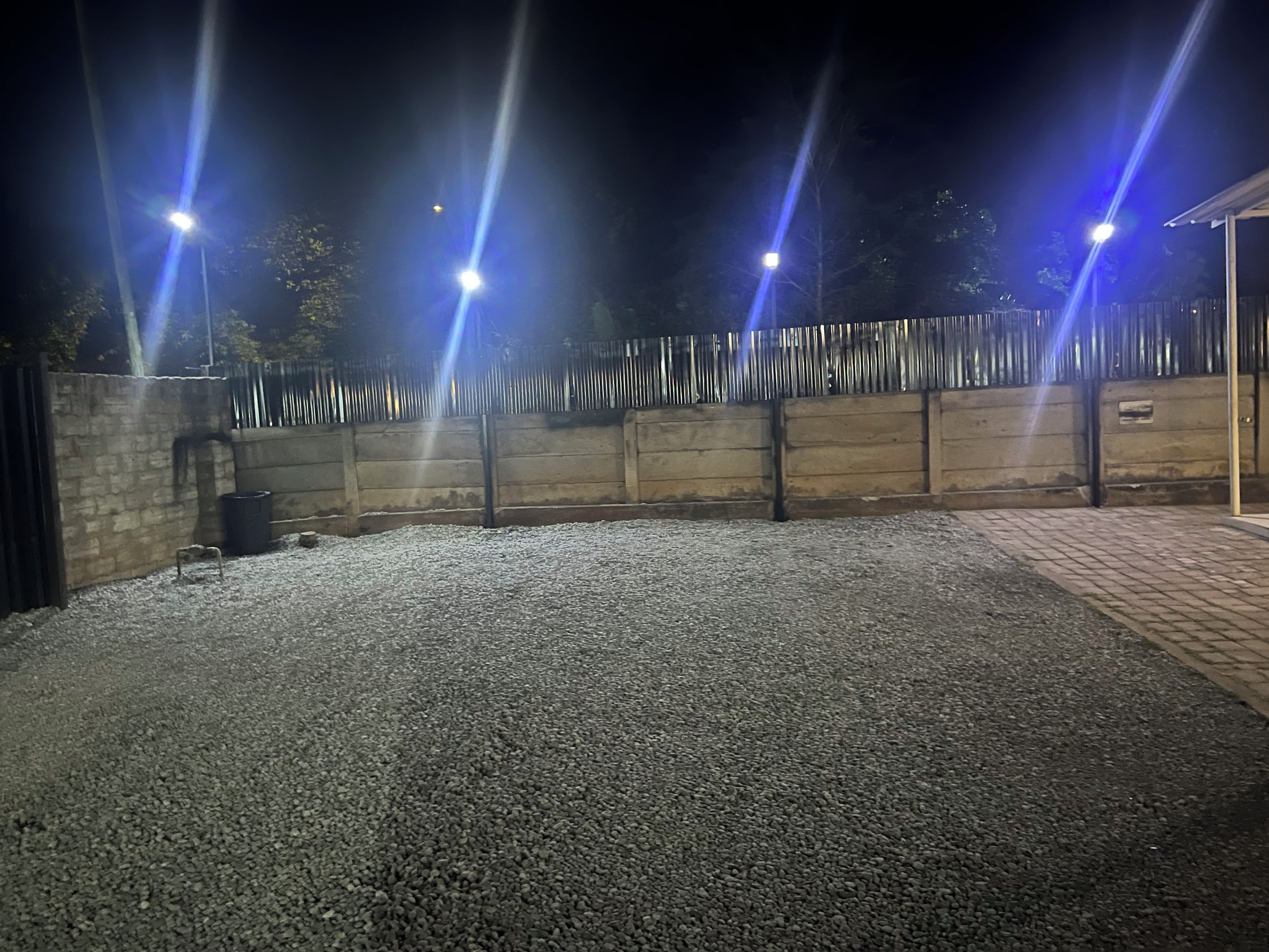 Un espacio al aire libre con suelo de grava, una valla de metal y varios postes de luz.
