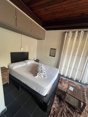 Un dormitorio con cama, mesita de noche, techo de madera, mueble de pared, ventana con cortinas y una alfombra.