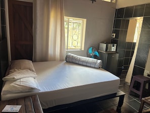 A single bed with a white mattress and a pillow, a small table with a sign, a fan, and a window with a view of greenery.