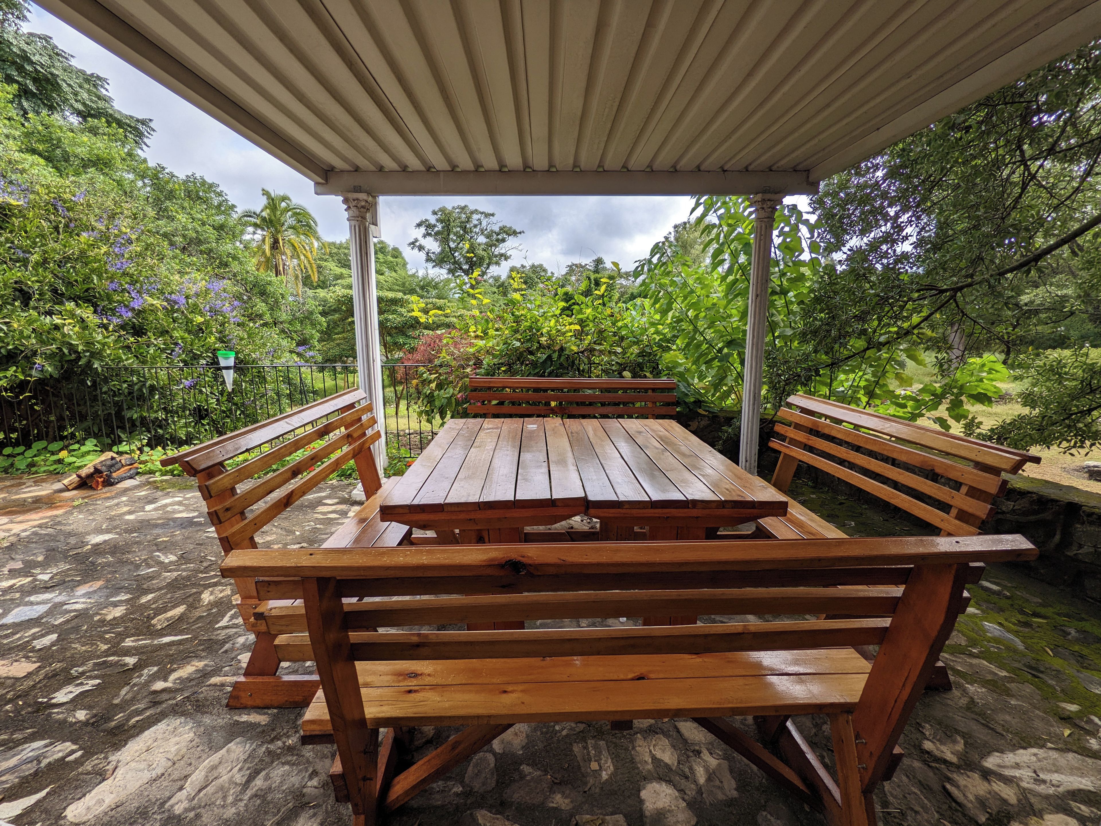 A covered outdoor dining area with a wooden table and benches, surrounded by lush greenery and a stone floor.