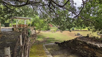 Un sendero de piedra conduce a un área de descanso cubierta con bancos, rodeada de abundante vegetación y un muro de piedra.