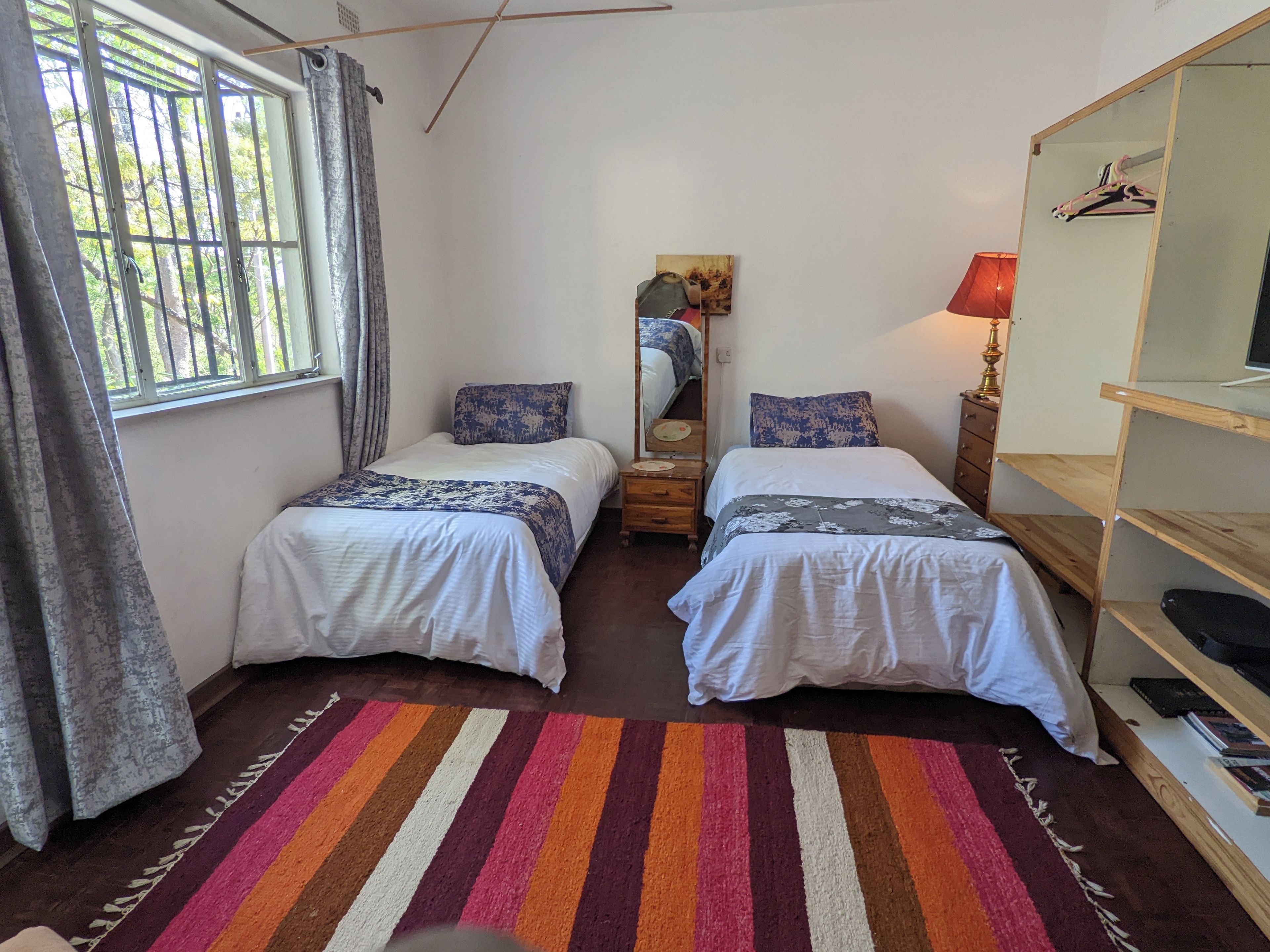 Een slaapkamer met twee bedden, een gestreept tapijt, een spiegel en een houten plank met boeken.