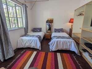A bedroom with two beds, a striped rug, a mirror, and a wooden shelf with books.