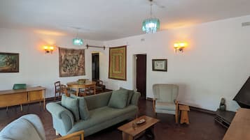 Una sala de estar con sofá, sillones, una mesa de centro, una mesa de comedor con sillas, una araña, luces de pared y obras de arte.