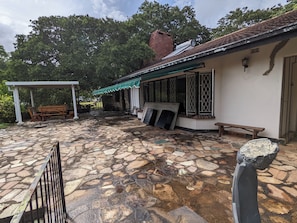 Un pati empedrat amb una pérgola i mobles de fusta, un edifici amb teulada i finestres, i una paret de pedra.