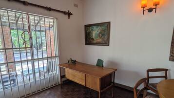 Une pièce avec un bureau en bois, une chaise, une fenêtre avec des barreaux en métal et un luminaire fixé au mur.