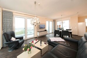 Ein modernes Wohnzimmer mit einer Couch, einem Couchtisch mit Blumen, einem Fernseher und einem Essbereich mit Stühlen und Tisch.