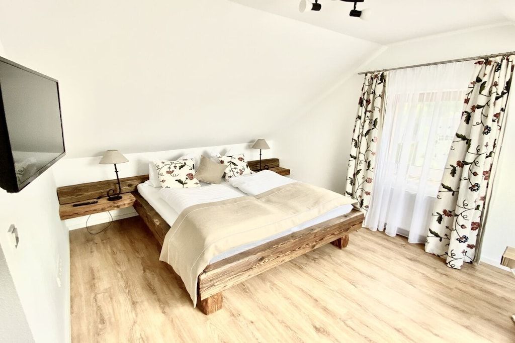 A bedroom with a wooden bed, a flat-screen TV mounted on the wall, a bedside table with a lamp, and a window with floral curtains.