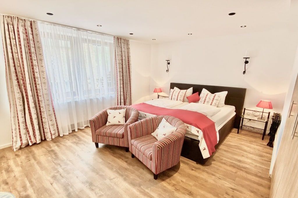 A hotel room with a bed, two armchairs, a bedside table with a lamp, and a window with curtains.
