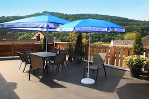 An outdoor patio with a table and chairs under blue umbrellas, surrounded by a wooden fence and potted plants.