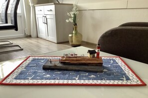 Um modelo de navio sobre uma mesa com toalha náutica, um vaso com flores e um armário ao fundo.