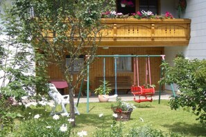 A garden with a swing set, a bench, and a wooden balcony with flowers. - Holiday apartment with 2 bedrooms (Roßhaupten)