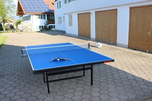 Une table de ping-pong installée à l’extérieur, avec un filet, une queue de billard et une boule, le tout placé sur une surface pavée avec un bâtiment en arrière-plan.