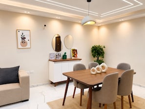 A modern dining area with a wooden table, grey upholstered chairs, a white cabinet, and a potted plant. - 3BRs AlAlson Institute Nasr City Fancy Apart (Cairo Governorate)
