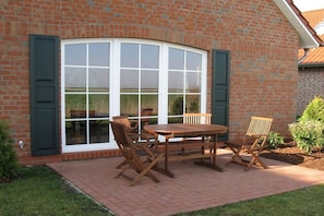 A brick building with a large window and a patio area with a wooden table and chairs. - Holiday home with 2 bedrooms (Greetsiel)