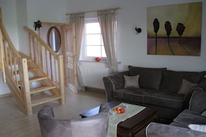 A living room with a wooden staircase, a grey sofa, a coffee table with fruit, and a painting on the wall. - Holiday home with 2 bedrooms (Greetsiel)