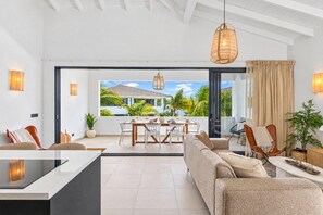 A modern living room with a sofa, dining table, and chairs, a large window with a view of a house and palm trees, and a hanging light fixture. - The Bank | Luxe Penthouses | 5 min Mambo&Jan thiel (Willemstad)